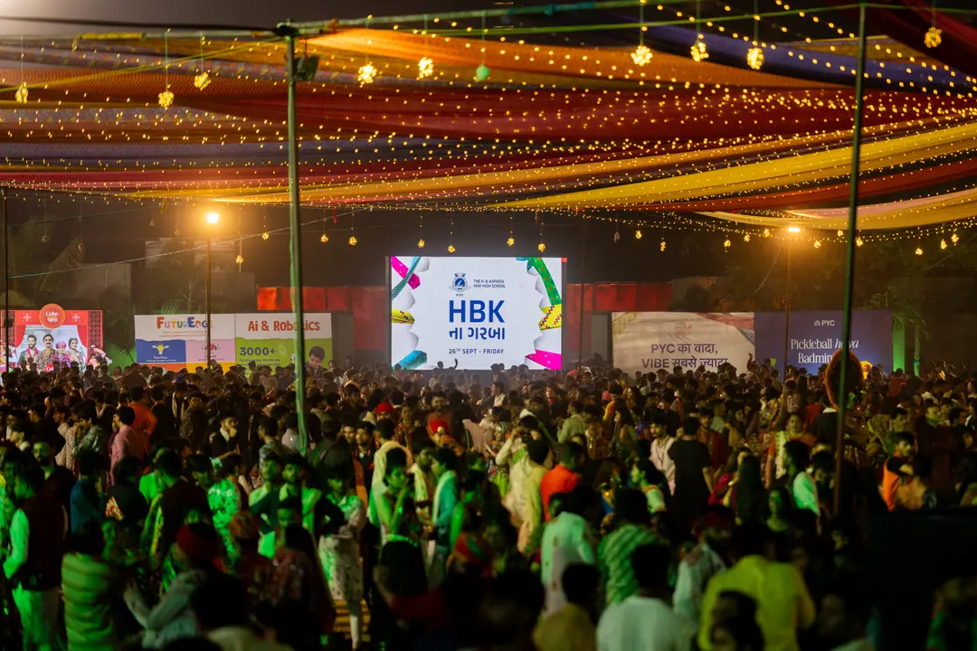 A packed garba ground under strings of canopy lights, the HBK ના ગરબા screen at the far end