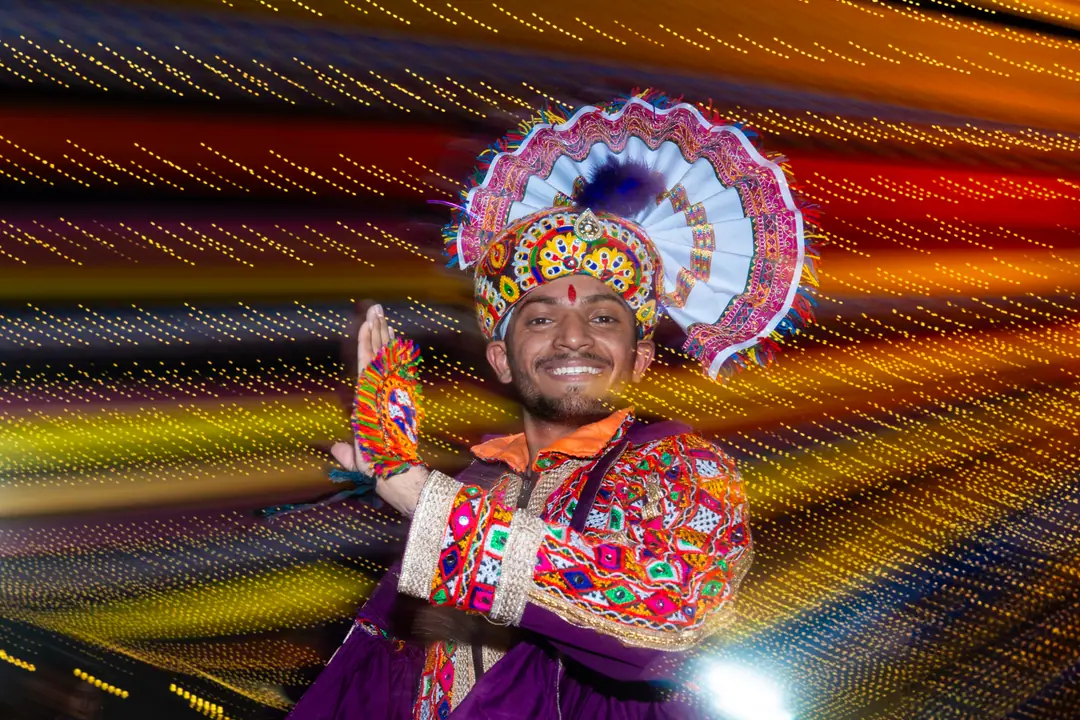 A grinning khelaiya in a peacock-fan headdress and a mirror-work kediyu, mid-garba under streaks of light