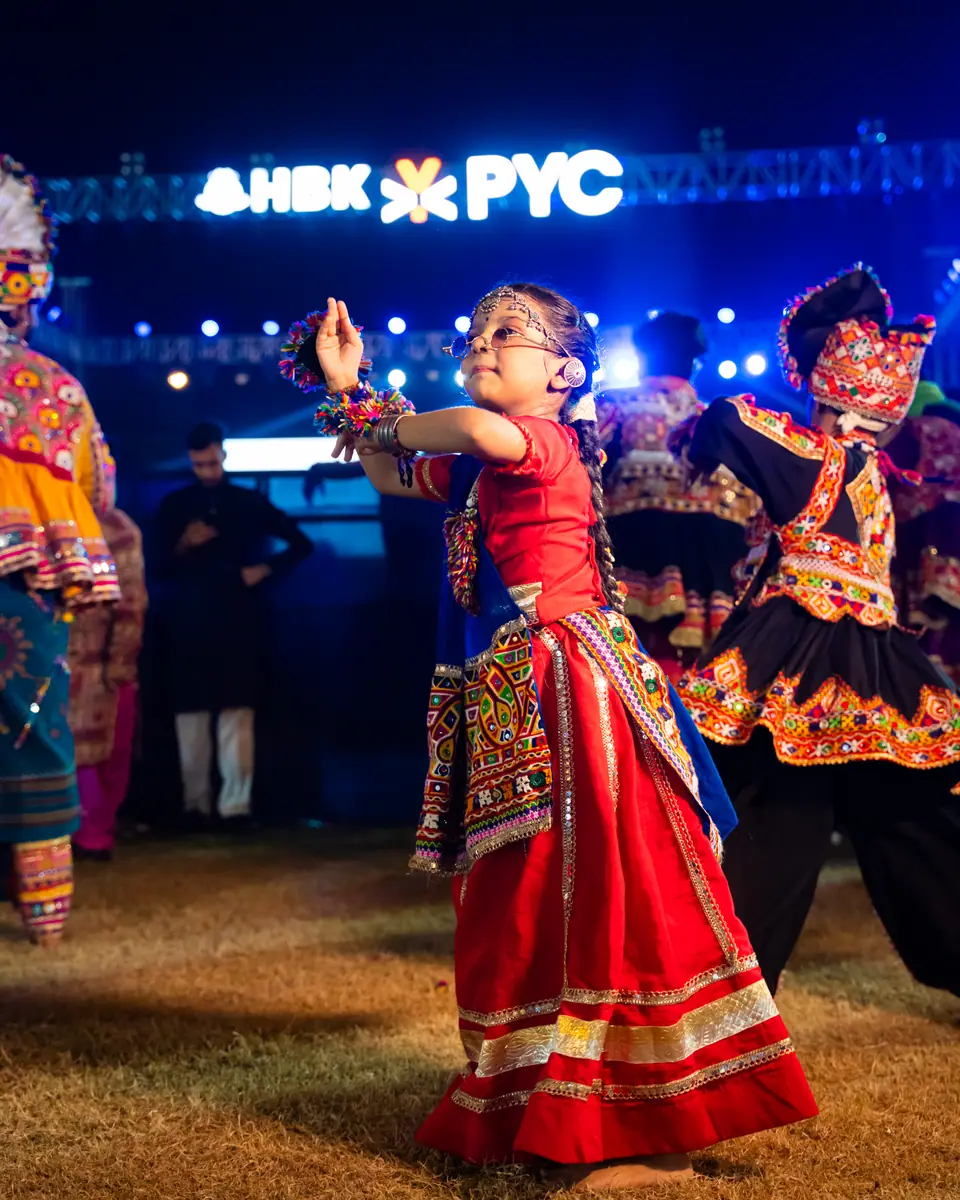 A girl in a red chaniya choli dancing among young khelaiyas, HBK ✕ PYC lit up behind them