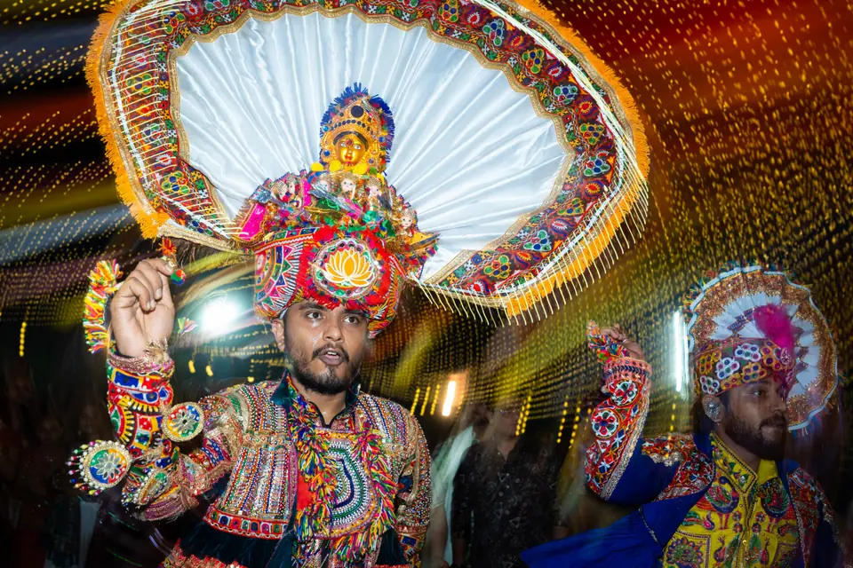 A khelaiya in a towering peacock-fan headdress, one hand raised, light streaking behind him